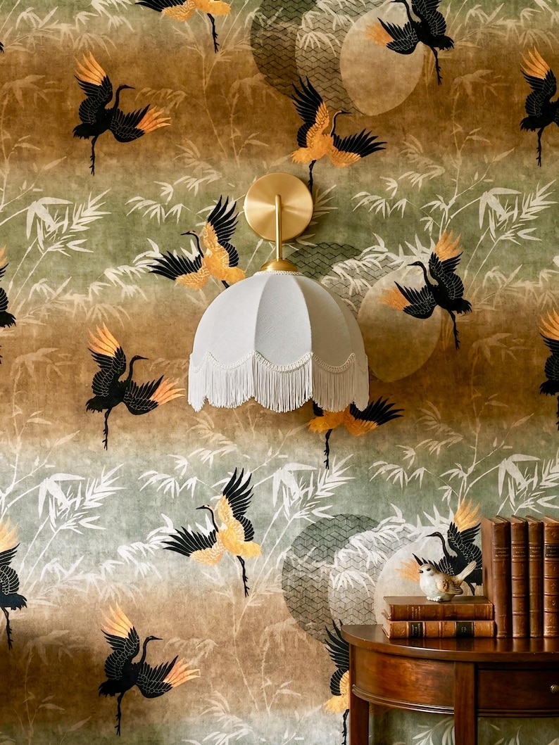 May include: A wall-mounted light fixture with a white lampshade and fringe is set against a wallpaper featuring a repeating pattern of black and gold cranes and bamboo leaves. A small wooden table holds books and a bird figurine.