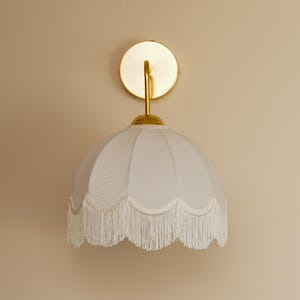 May include: A white wall sconce with a scalloped lampshade and fringe trim. The sconce has a gold-colored circular backplate and a curved arm. The lampshade is a soft, off-white color.
