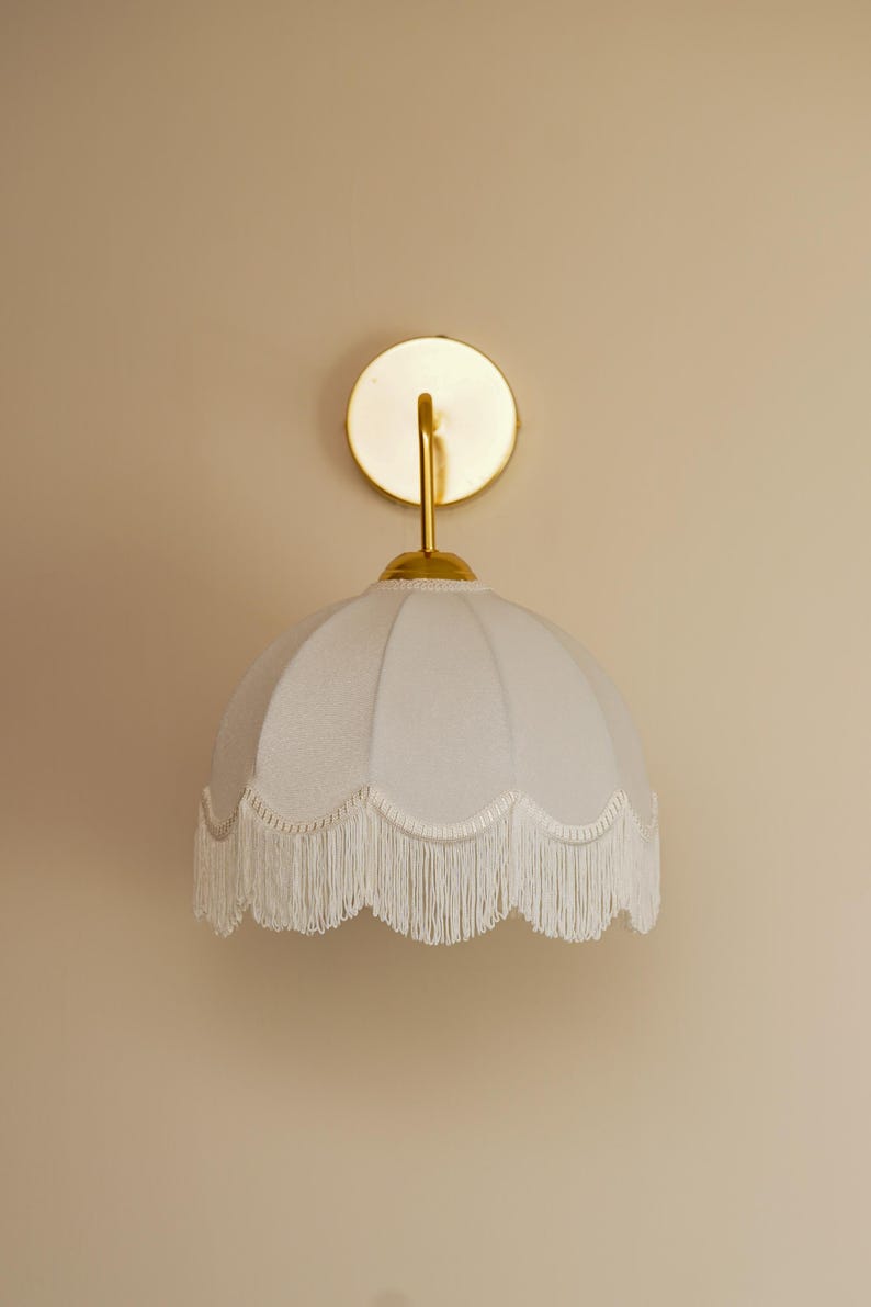 May include: A white wall sconce with a scalloped lampshade and fringe trim. The sconce has a gold-colored circular backplate and a curved arm. The lampshade is a soft, off-white color.