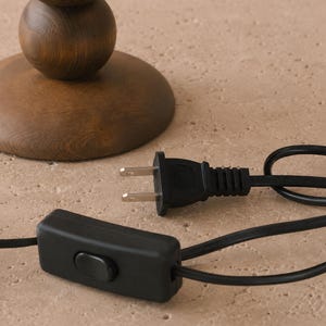 May include: A dark brown wooden lamp base with a stacked spherical design, featuring a black electrical cord with a two-prong plug and a rectangular inline rocker switch. The cord is black and extends from the base of the wooden lamp structure.