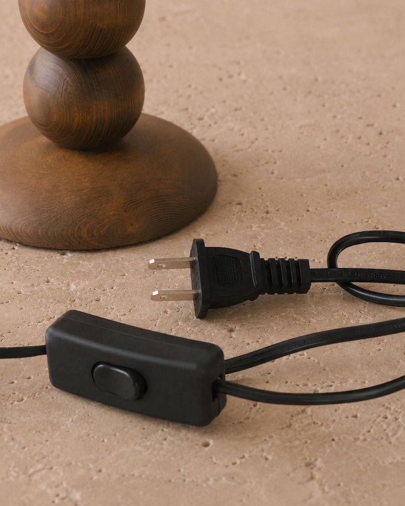 May include: A dark brown wooden lamp base with a stacked spherical design, featuring a black electrical cord with a two-prong plug and a rectangular inline rocker switch. The cord is black and extends from the base of the wooden lamp structure.