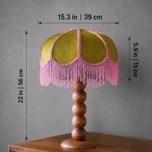 May include: A table lamp with a dome-shaped olive green velvet shade featuring pink braided trim and long pink fringe. The base consists of four stacked wooden spheres on a circular stand. Dimensions are 15.3 inches wide, 5.9 inches shade height, 22 inches total height.