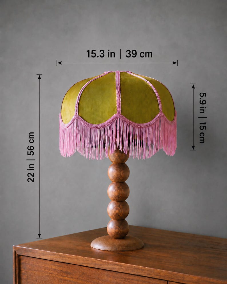May include: A table lamp with a dome-shaped olive green velvet shade featuring pink braided trim and long pink fringe. The base consists of four stacked wooden spheres on a circular stand. Dimensions are 15.3 inches wide, 5.9 inches shade height, 22 inches total height.