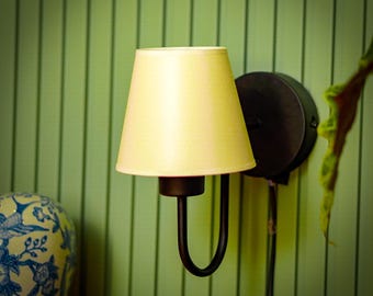 May include: A wall-mounted lamp with a pale yellow lampshade and a dark metal base. The lamp has a curved arm and a circular mounting plate. The background is a light green wall with vertical lines. A decorative ceramic object is visible in the lower left corner.