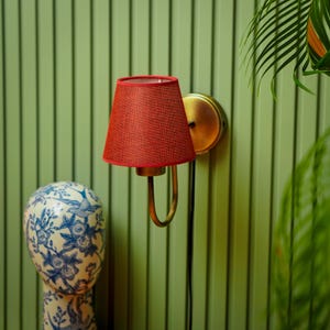 Puede incluir: Una pantalla de lámpara de tela roja con una aplique de pared de metal dorado. La pantalla está unida a la aplique con un brazo de metal dorado. La aplique está montada en una pared verde.