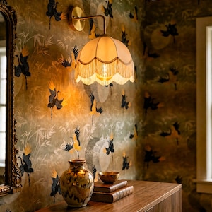 May include: A vintage-style wall sconce with a fringed lampshade illuminates a room with patterned wallpaper. The wallpaper features a crane design in black and yellow against a muted green background. A wooden cabinet holds a vase, books, and a bowl.