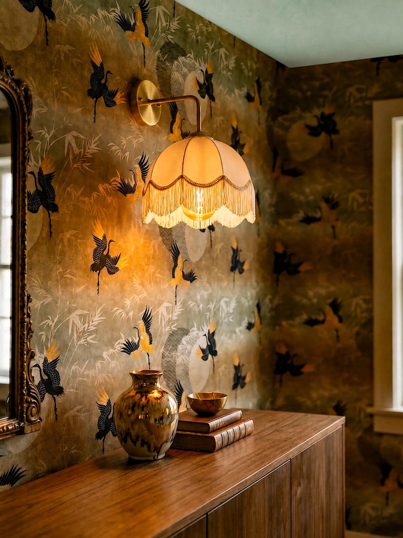 May include: A vintage-style wall sconce with a fringed lampshade illuminates a room with patterned wallpaper. The wallpaper features a crane design in black and yellow against a muted green background. A wooden cabinet holds a vase, books, and a bowl.