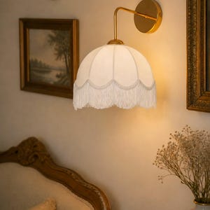 May include: A wall-mounted lamp with a white, scalloped shade and fringe trim. The lamp has a gold-colored arm and base. The image also shows framed artwork and a decorative vase, suggesting a vintage or antique aesthetic.