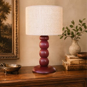 May include: A table lamp featuring a cylindrical, textured off-white fabric lampshade and a base composed of four stacked, smooth, deep burgundy spheres resting on a circular pedestal of the same dark red colour and matte finish.