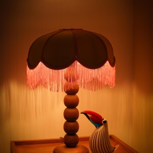 May include: A table lamp with a stacked wooden sphere base and a scalloped fabric shade featuring long red fringe trim. Beside the lamp sits a glass toucan figurine with black and white stripes and a red, blue, and black beak, plus a small square tile.