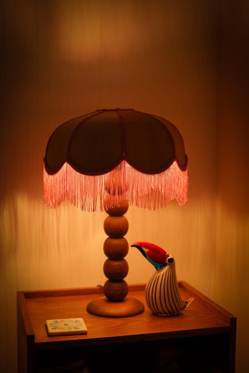May include: A table lamp with a stacked wooden sphere base and a scalloped fabric shade featuring long red fringe trim. Beside the lamp sits a glass toucan figurine with black and white stripes and a red, blue, and black beak, plus a small square tile.