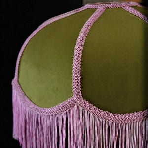 May include: A dome-shaped lampshade covered in olive green velvet fabric. The shade features decorative light pink braided trim along the seams and a long, matching light pink fringe hanging from the scalloped bottom edge of the lampshade.