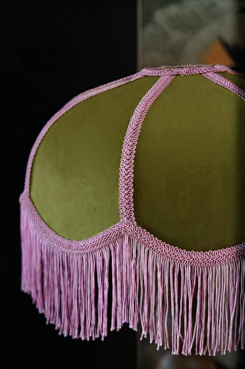 May include: A dome-shaped lampshade covered in olive green velvet fabric. The shade features decorative light pink braided trim along the seams and a long, matching light pink fringe hanging from the scalloped bottom edge of the lampshade.