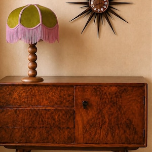 May include: A wooden sideboard with a burl wood veneer finish, featuring a table lamp with a green velvet shade and pink fringe, and a dark brown starburst wall clock with thin radiating spikes and a circular face with white numerals.