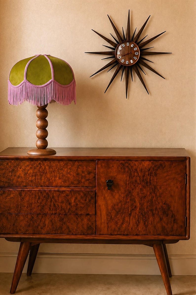 May include: A wooden sideboard with a burl wood veneer finish, featuring a table lamp with a green velvet shade and pink fringe, and a dark brown starburst wall clock with thin radiating spikes and a circular face with white numerals.