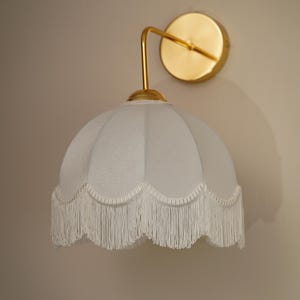 May include: A wall-mounted light fixture with a gold-colored arm and circular backplate. The light has a pale blue, scalloped shade with white fringe trim. The fixture is mounted on a neutral-colored wall.
