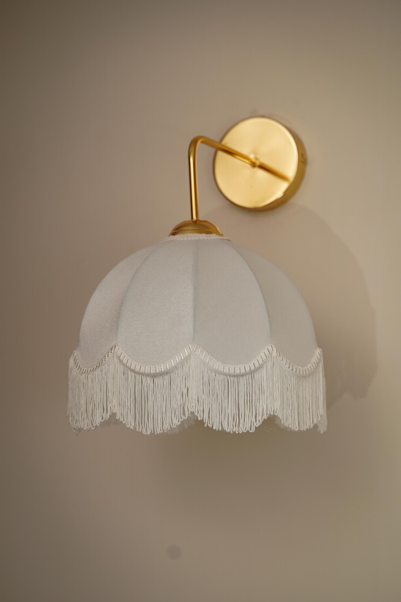 May include: A wall-mounted light fixture with a gold-colored arm and circular backplate. The light has a pale blue, scalloped shade with white fringe trim. The fixture is mounted on a neutral-colored wall.