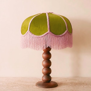 May include: A table lamp featuring a dome-shaped lampshade covered in olive green fabric with pink braided trim and long pink fringe. The base is constructed from a vertical stack of five smooth, round, dark brown wooden spheres on a circular stand.