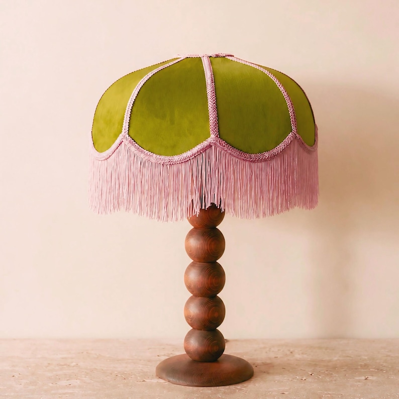 May include: A table lamp featuring a dome-shaped lampshade covered in olive green fabric with pink braided trim and long pink fringe. The base is constructed from a vertical stack of five smooth, round, dark brown wooden spheres on a circular stand.