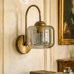 May include: A wall-mounted lamp featuring a curved brass arm and a circular brass base. The fixture includes a rounded, transparent glass shade with a smoke-tinted finish, housing a single light bulb visible within the clear glass structure.