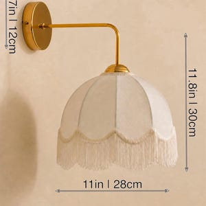 May include: A gold-toned wall sconce with a cream-colored, fringed lampshade. The sconce is 13.4 inches (34 cm) wide, 4.7 inches (12 cm) high, and the shade is 11 inches (28 cm) wide and 11.8 inches (30 cm) high. The fixture has a classic, elegant design.