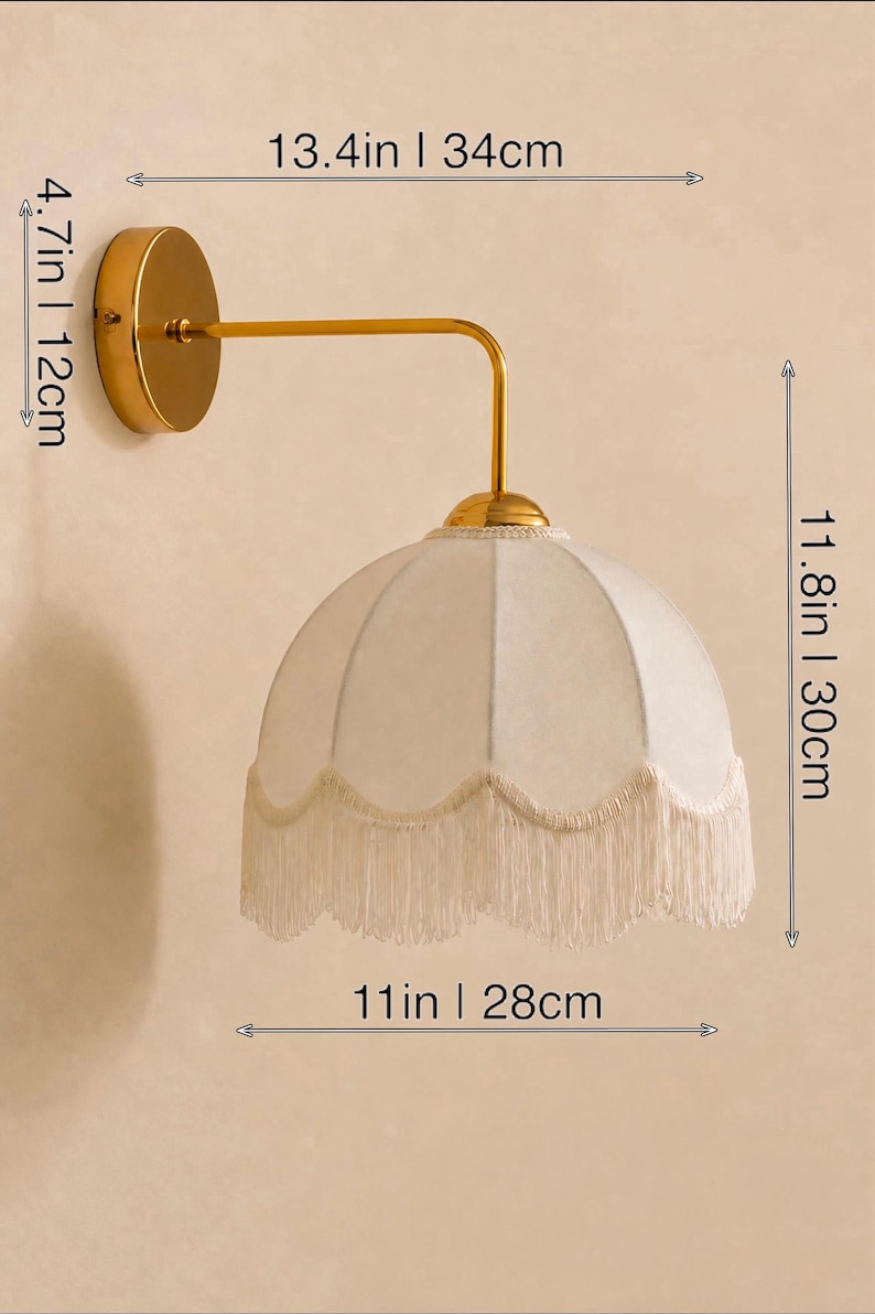 May include: A gold-toned wall sconce with a cream-colored, fringed lampshade. The sconce is 13.4 inches (34 cm) wide, 4.7 inches (12 cm) high, and the shade is 11 inches (28 cm) wide and 11.8 inches (30 cm) high. The fixture has a classic, elegant design.