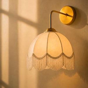 May include: A wall-mounted lamp with a cream-colored, fringed shade. The lamp has a brass-colored arm and a circular base. The shade is dome-shaped and has a scalloped edge with hanging fringe. The lamp is mounted on a light-colored wall.