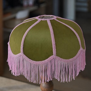 May include: A dome-shaped lampshade featuring a smooth olive green velvet fabric. The shade is accented with decorative light pink braided trim along the seams and edges, finished with a long, flowing light pink fringe hanging from the bottom.