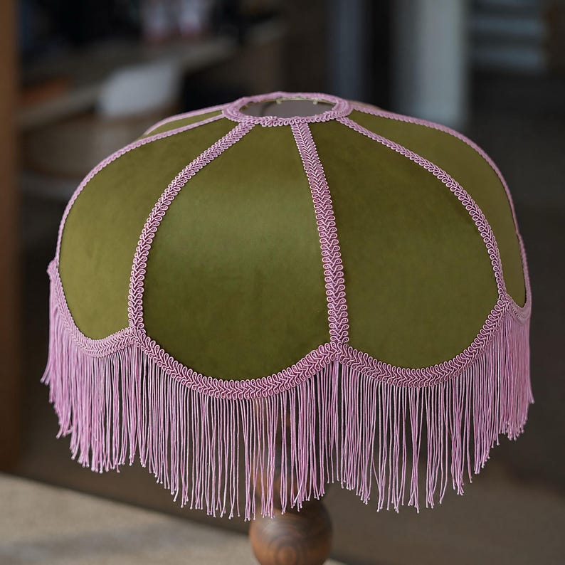 May include: A dome-shaped lampshade featuring a smooth olive green velvet fabric. The shade is accented with decorative light pink braided trim along the seams and edges, finished with a long, flowing light pink fringe hanging from the bottom.