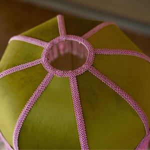 May include: A hexagonal lampshade featuring olive green velvet fabric panels. The edges and the circular top opening are finished with decorative pink braided trim, creating a structured geometric design with a soft, textured appearance.