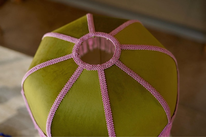 May include: A hexagonal lampshade featuring olive green velvet fabric panels. The edges and the circular top opening are finished with decorative pink braided trim, creating a structured geometric design with a soft, textured appearance.