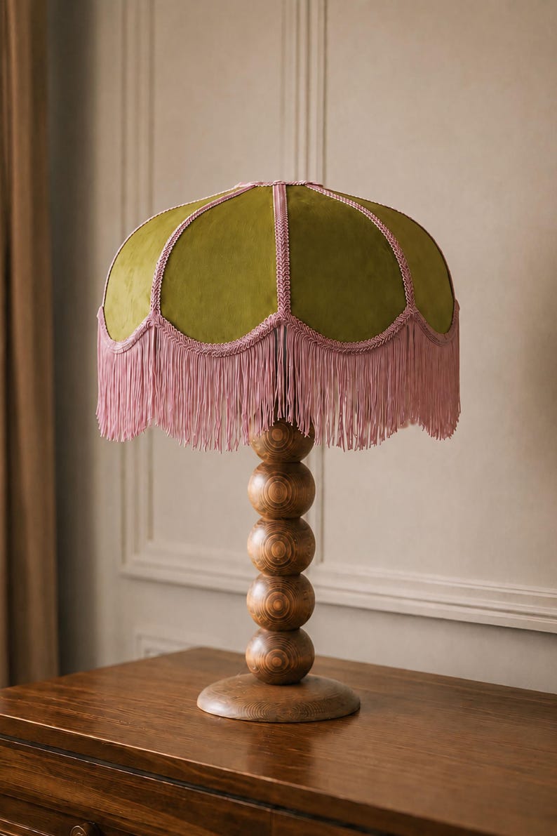 A table lamp featuring a dome-shaped olive green velvet lampshade with pink braided trim and a long pink fringe. The lamp base is constructed from  stacked wooden spheres resting on a circular wooden stand with a visible grain.