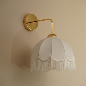 May include: A wall-mounted light fixture with a gold finish. The fixture features a circular base, a curved arm, and a white lampshade with a scalloped edge and fringe. The fixture is designed for indoor use.
