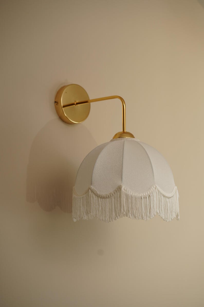 May include: A wall-mounted light fixture with a gold finish. The fixture features a circular base, a curved arm, and a white lampshade with a scalloped edge and fringe. The fixture is designed for indoor use.