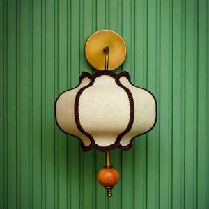 Puede incluir: Un aplique de pared con una pantalla de tela color crema festoneada con ribete marrón oscuro. La lámpara incluye una placa de pared circular de latón, un brazo de latón curvado y un adorno de esfera de madera naranja en la parte inferior.