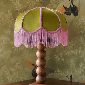 May include: A table lamp featuring a dome-shaped olive green velvet shade with pink braided trim and long pink fringe. The base is constructed from five stacked wooden spheres of decreasing size, supported by a circular wooden pedestal stand.