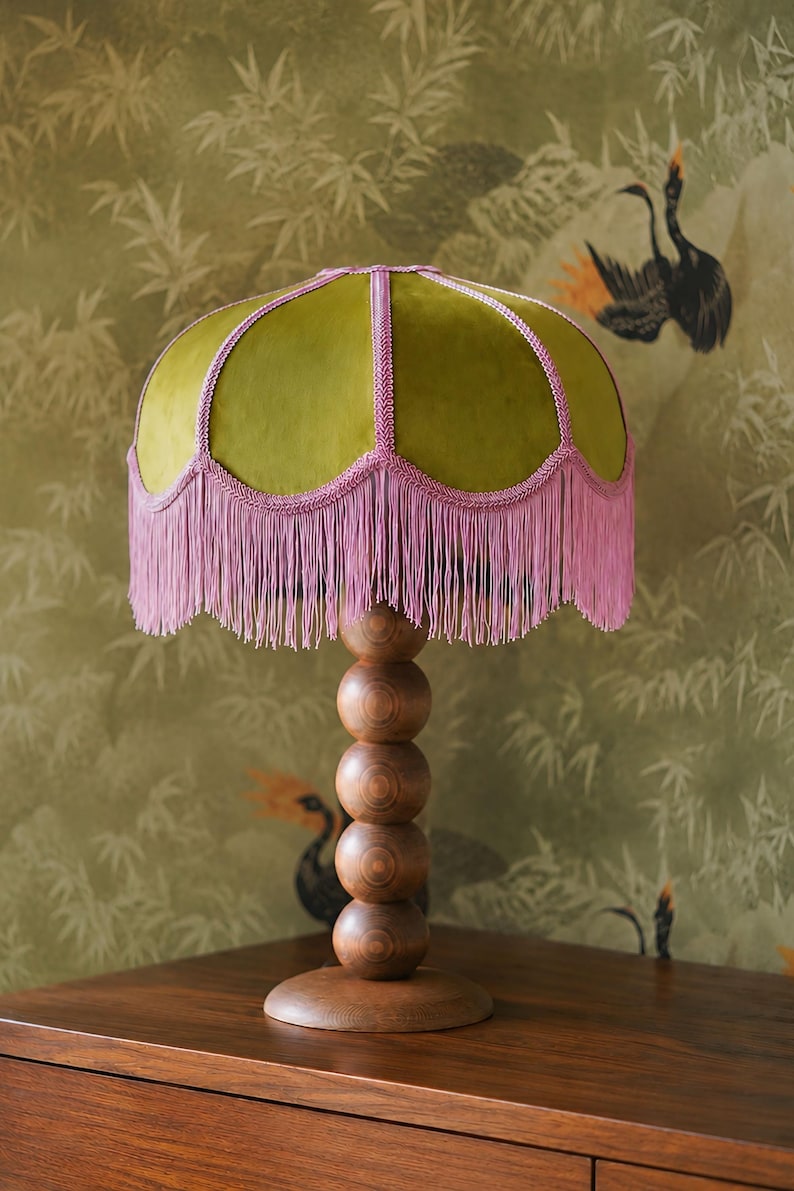 May include: A table lamp featuring a dome-shaped olive green velvet shade with pink braided trim and long pink fringe. The base is constructed from five stacked wooden spheres of decreasing size, supported by a circular wooden pedestal stand.