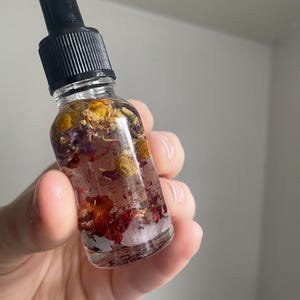 May include: A clear glass bottle with a black dropper top, filled with a pinkish liquid and dried flowers. The bottle is held in a hand against a plain background. The flowers are yellow, red, and purple.