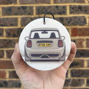 May include: A white round car air freshener featuring a grey Mini Cooper. The car is facing away from the viewer and has the number plate "FF56 COP".
