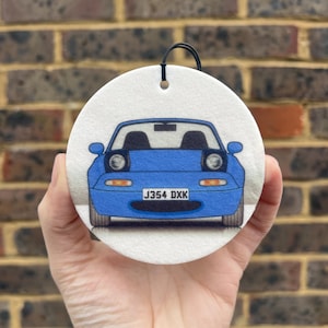 May include: A circular white air freshener featuring a front-facing illustration of a blue convertible sports car with pop-up headlights. The car has a license plate that reads J354 DXK. A black elastic string is attached to the top of the ornament.