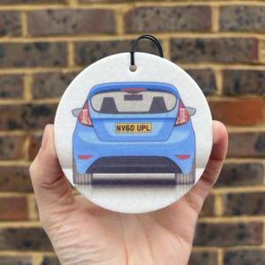 May include: A white round car air freshener with a blue Ford Fiesta printed on it. The car is facing away from the viewer and the license plate reads "NV60 UPL".