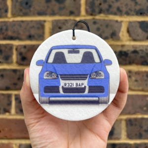 May include: A circular car air freshener with a blue car illustration. The car has headlights and a registration plate reading "R321 BAP". A black string is attached for hanging. The background is a brick wall.