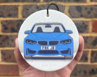 May include: A round, white car air freshener featuring a blue sports car illustration. The car has a black roof and the license plate reads "F8X JB". A black string is attached for hanging.