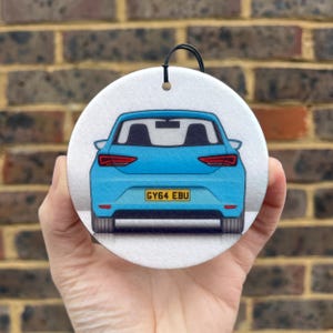 May include: A circular white air freshener featuring a graphic illustration of the rear view of a bright blue hatchback car. The car has red taillights, black tyres, and a yellow number plate with the black text GY64 EBU printed on it.