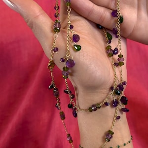 Könnte beinhalten: Goldfarbene Halskette mit mehreren Strängen, verziert mit lila und grünen Edelsteinen. Die Halskette hat ein mehrschichtiges, zartes Design, perfekt, um jedem Outfit einen Hauch von Eleganz zu verleihen. Die Edelsteine sind in verschiedenen Formen und Größen.