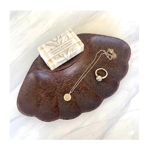 May include: A dark brown wooden tray with a scalloped edge holds a rectangular bar of Aromatic Botanicals Sandalwood & Myrrh soap wrapped in paper, a gold chain necklace with a starburst pendant, and a gold ring featuring a white pearl stone.