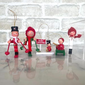 May include: A group of five wooden Christmas tree decorations. The ornaments feature skiers, a soldier, and a child on a sled. One ornament has a sign that reads "MERRY CHRISTMAS". The ornaments are painted in red, white, black, and green.
