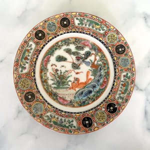 May include: A decorative plate with a detailed, colourful design. The plate features a central scene with a bird, a fox, and foliage, surrounded by a border of coins and floral patterns. The colour palette includes red, green, blue, and gold.