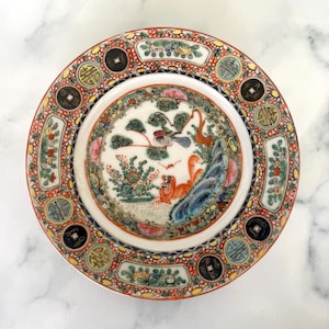 May include: A round, decorative plate with a detailed, colourful design. The plate features a central image of birds, a tiger, and foliage, surrounded by a red border with floral and coin-like motifs. The plate has a white interior and an ornate pattern.