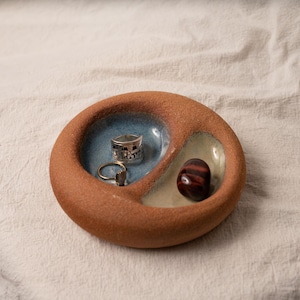 May include: A round, terracotta-colored ceramic dish with a textured exterior and two interior compartments. One compartment is glazed in blue and holds two silver-toned rings, while the other is glazed in cream and holds a polished red-brown stone.